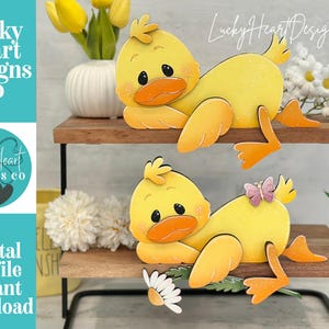May include: Two wooden yellow duck decorations with orange beaks and feet are displayed on a two-tiered wooden shelf. The ducks are lying down. The image includes the text "Lucky Heart Designs Co" and "Digital cut file instant download".