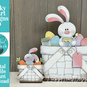 May include: Wooden Easter basket decorations featuring bunnies and painted eggs. The larger basket has a bunny with a paintbrush and the text "HOPPY EASTER" on a tag. The smaller basket has a bunny with carrots. The text "Lucky Heart Designs Co" is also visible.