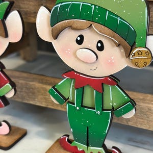May include: A wooden Christmas elf decoration. The elf has a green hat and outfit with red accents. The elf has a rosy complexion, black eyes, and a small bell on its hat. The decoration is mounted on a small wooden base.