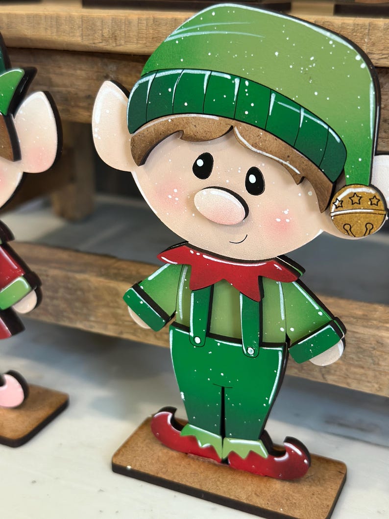 May include: A wooden Christmas elf decoration. The elf has a green hat and outfit with red accents. The elf has a rosy complexion, black eyes, and a small bell on its hat. The decoration is mounted on a small wooden base.