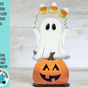 Puede incluir: Un fantasma blanco sosteniendo una cuerda de maíz dulce sobre una calabaza tallada en forma de cara. La calabaza está sobre una base de madera. El fantasma y la calabaza están pintados en un estilo caprichoso.
