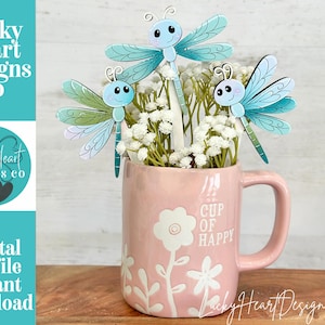 May include: A pink mug with the words "CUP OF HAPPY" and floral designs holds white flowers and three dragonfly cutouts. The dragonflies are light blue and green with smiling faces. The image also includes the text "Lucky Heart Designs Co."