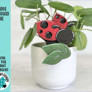 Op de afbeelding: Een rode en zwarte lieveheersbeestje mokwarmer met een glimlachend gezicht. Het lieveheersbeestje zit op een groen blad in een witte pot.