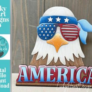 May include: A patriotic wooden eagle decoration with sunglasses and a headband, featuring the American flag design. The word "AMERICA" is displayed in red and blue. The design is from Lucky Heart Designs Co.