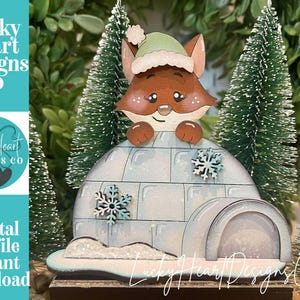 Könnte beinhalten: Ein hölzernes Fuchsornament mit grünem Hut, das über einem hellblauen Iglu mit Schneeflocken-Akzenten hervorlugt. Das Ornament trägt den Text "Lucky Heart Designs Co" und "Digital cut file instant download".