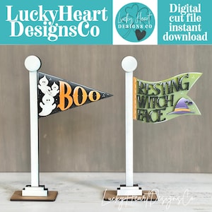 May include: Two white wooden flag stands with decorative pennants. The left pennant is black with white ghosts and the orange text BOO. The right pennant is light green with the black text RESTING WITCH FACE and a purple and yellow witch hat icon.