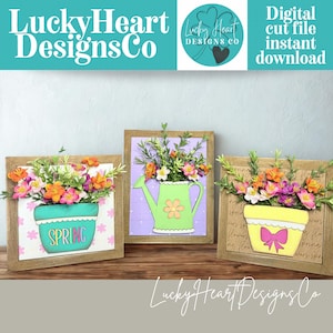 May include: Three framed floral art pieces. Each features a wooden frame with a colorful flower arrangement. One has a turquoise pot with the word "SPRING". Another has a green watering can. The third has a yellow pot with a pink bow. The text "Lucky Heart Designs Co" is visible.
