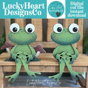 May include: Two green frog cutouts with large white eyes and dark pupils. The frogs have speckled skin, smiling mouths, and are positioned in a seated pose. One frog is flanked by two small brown mushrooms. Text reads LuckyHeartDesignsCo.