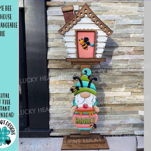 Könnte beinhalten: Ein Vogelhaus im Gnome-Stil mit Bienenmotiv. Das Vogelhaus hat einen weißen Körper, ein braunes Dach und eine rosa Tür mit einer Bienenillustration. Eine Zwergfigur hält einen Honigtopf. Der Text "HONEY" ist sichtbar.