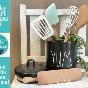 May include: A black ceramic canister with the word YUM printed in white, containing wooden kitchen utensil cutouts including a spatula, a whisk, and a spoon. A wooden rolling pin lies in front of the canister next to a black lid.