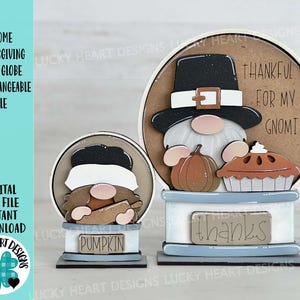 May include: Two wooden gnome snow globe ornaments. The larger snow globe features a gnome with a black hat and a white beard holding a pumpkin and a pie. The smaller snow globe features a gnome with a black hat and a white beard holding a pumpkin. The larger snow globe has the text "Thankful for my Gnomies" and "Thanks" on it.