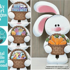 May include: A white wooden Easter bunny decoration holding a brown basket filled with orange carrots. The bunny has pink inner ears and a pink nose. The text "Lucky Heart Designs Co" is visible.