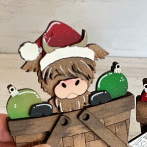K&ouml;nnte beinhalten: Ein h&ouml;lzerner Weihnachtsbaumschmuck mit einer schottischen Hochlandkuh, die einen roten Weihnachtsmann-Hut tr&auml;gt und gr&uuml;ne Kugeln in einem braunen Korb h&auml;lt.