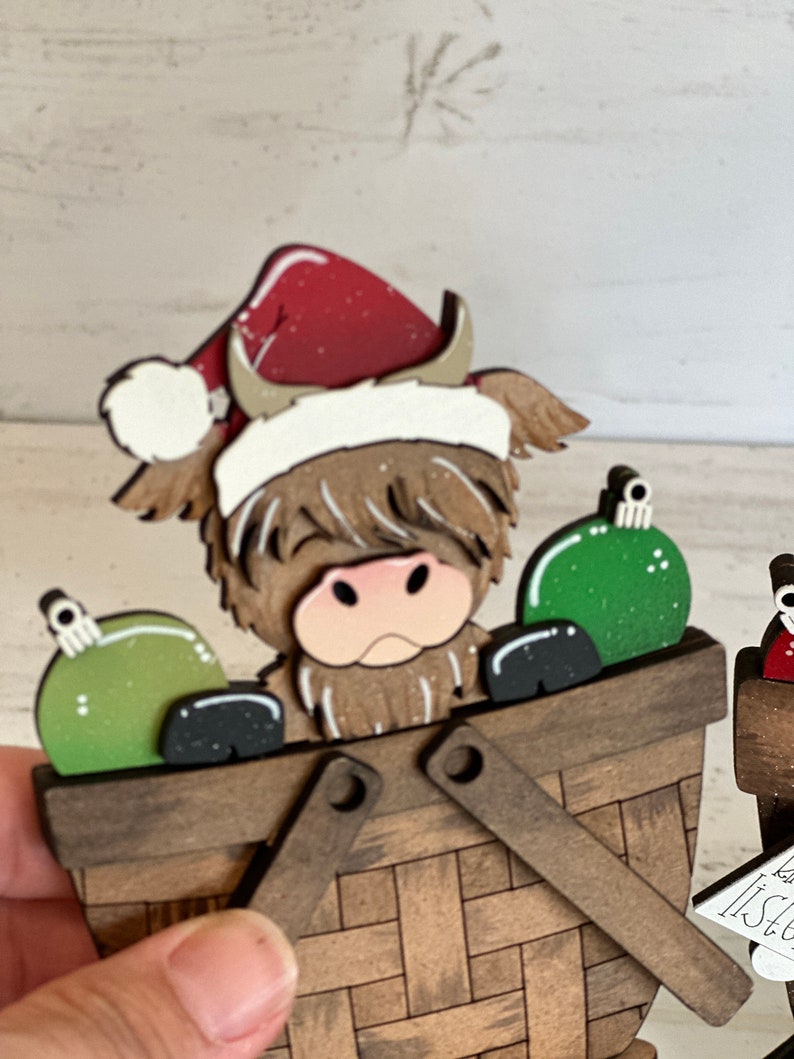 K&ouml;nnte beinhalten: Ein h&ouml;lzerner Weihnachtsbaumschmuck mit einer schottischen Hochlandkuh, die einen roten Weihnachtsmann-Hut tr&auml;gt und gr&uuml;ne Kugeln in einem braunen Korb h&auml;lt.