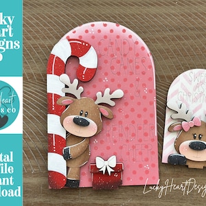 May include: Two arched wooden signs featuring cartoon reindeer. The larger sign has a red and white candy cane, a red gift box with a white bow, and the text Candy Canes & Reindeer Cheer. The smaller sign features a reindeer with a pink bow and the text Sweet As Christmas.