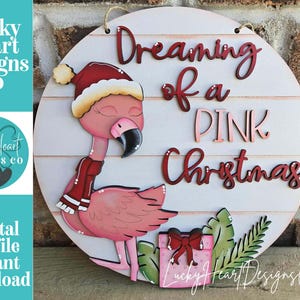 Op de afbeelding: Ronde houten bord met een flamingo met een kerstmuts en sjaal, met de tekst "Dreaming of a PINK Christmas". De flamingo is roze met een zwarte snavel. Een geschenkdoos en groen zijn ook inbegrepen.