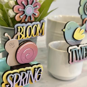 May include: A set of four spring-themed wooden decorations with glitter accents. The decorations feature a flower, a snail, a bird, and the word "Spring".