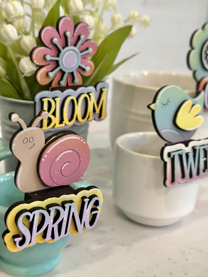 May include: A set of four spring-themed wooden decorations with glitter accents. The decorations feature a flower, a snail, a bird, and the word "Spring".