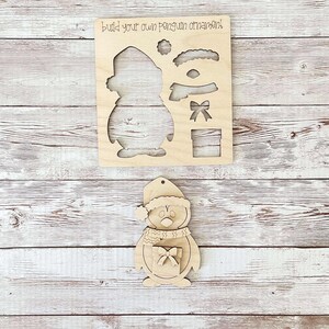 May include: Wooden penguin ornament craft kit. The kit includes a penguin shape with a hat, scarf, eyes, beak, and a gift box. The text "Build your own penguin ornament" is printed on the wood.