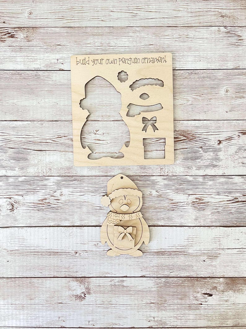 May include: Wooden penguin ornament craft kit. The kit includes a penguin shape with a hat, scarf, eyes, beak, and a gift box. The text "Build your own penguin ornament" is printed on the wood.