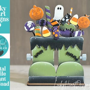 May include: A decorative wooden display featuring two green boots with black stitched details and silver bolts, topped with an assortment of Halloween-themed cutouts including a ghost, pumpkins, candy corn, lollipops, and a purple candy-coated apple.
