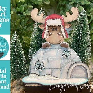 May include: A wooden Christmas decoration featuring a cartoon moose wearing a red hat and sitting on an igloo. The igloo has snowflake accents. The text "Lucky Heart Designs Co" and "Digital cut file instant download" are visible.