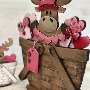 May include: A wooden moose figurine wearing a pink hat and holding a basket filled with pink hearts, candy, and a gift tag. The moose is standing in a brown wooden basket with a white and pink heart design.