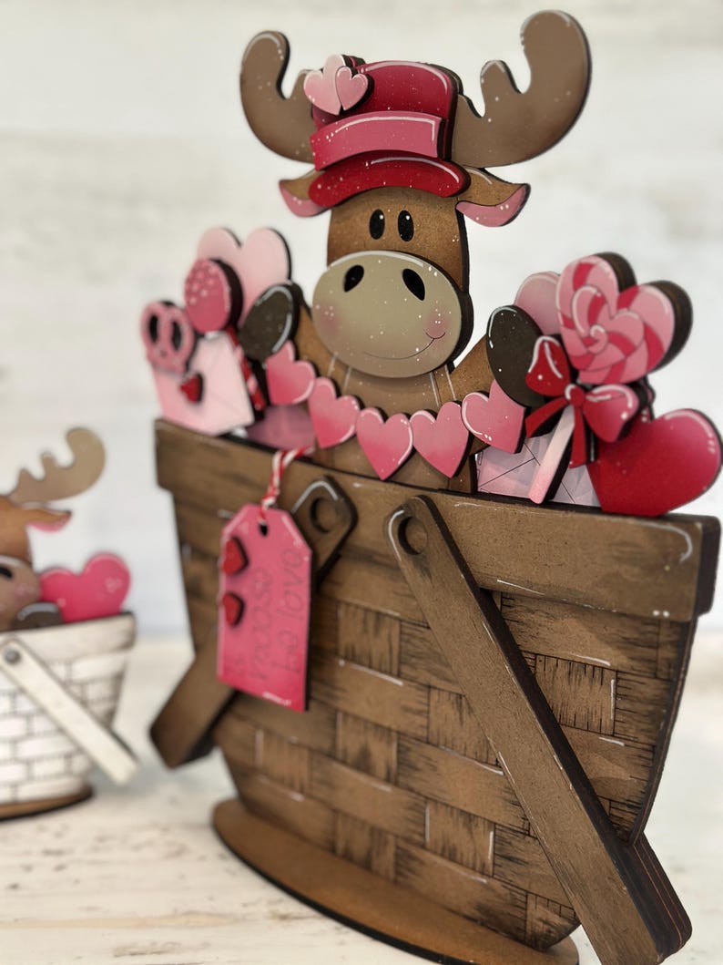 May include: A wooden moose figurine wearing a pink hat and holding a basket filled with pink hearts, candy, and a gift tag. The moose is standing in a brown wooden basket with a white and pink heart design.