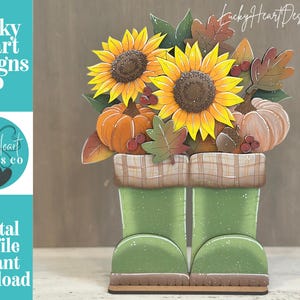 May include: A decorative wooden cutout featuring two green rain boots filled with yellow sunflowers, orange and white pumpkins, brown acorns, and autumn leaves. The boots have a tan plaid cuff. The text Lucky Heart Designs Co is visible.