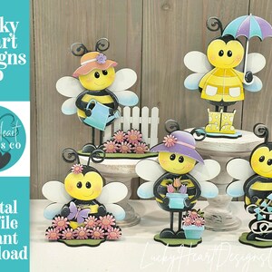 May include: A collection of whimsical bee figurines, each with unique accessories like hats, umbrellas, and watering cans. The bees are yellow and black, with white wings, and are set against a wooden background. The image includes the text "Lucky Heart Designs Co."