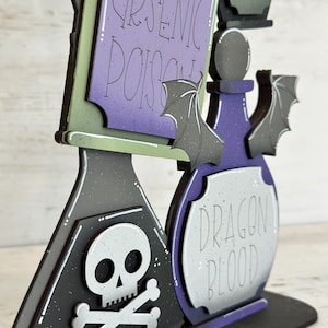 May include: A Halloween-themed wooden decoration featuring three potion bottles and a sign. The sign reads "Arsenic Poison" and "Venom". The bottles are labeled "Dragon Blood". The color scheme includes black, purple, green, and gray with bat and skull accents.