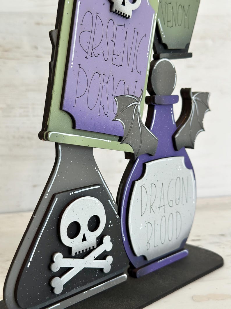 May include: A Halloween-themed wooden decoration featuring three potion bottles and a sign. The sign reads "Arsenic Poison" and "Venom". The bottles are labeled "Dragon Blood". The color scheme includes black, purple, green, and gray with bat and skull accents.