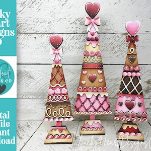 Puede incluir: Tres árboles decorativos de madera para el Día de San Valentín. Cada árbol tiene un diseño diferente, con colores rosa, marrón y blanco. Los árboles tienen adornos en forma de corazón y están decorados con corazones y lazos. El texto "Lucky Heart Designs Co" es visible.