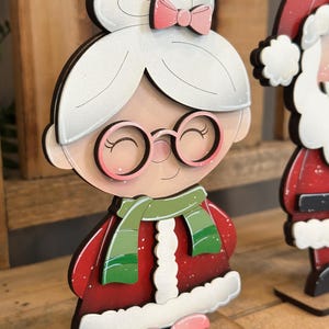 May include: Wooden Mrs. Claus Christmas decoration. The figure has a white hairstyle with a pink bow, round glasses, and a green scarf. She wears a red coat with white trim and pink shoes. The figure is on a wooden base.