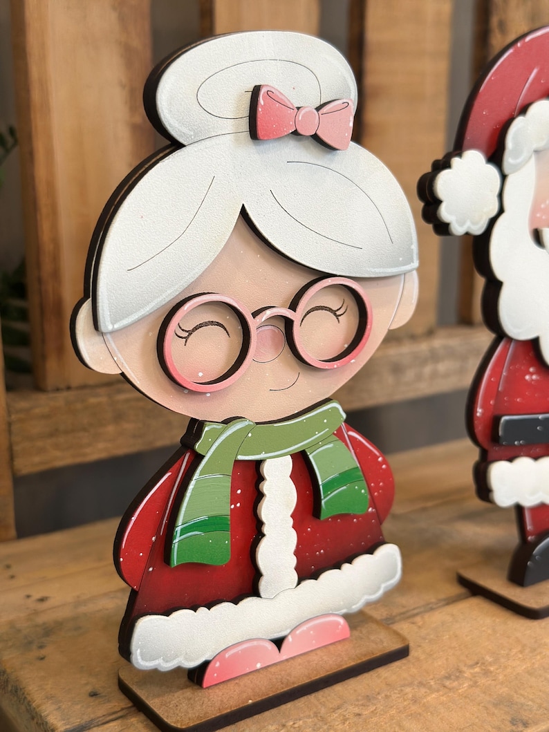 May include: Wooden Mrs. Claus Christmas decoration. The figure has a white hairstyle with a pink bow, round glasses, and a green scarf. She wears a red coat with white trim and pink shoes. The figure is on a wooden base.