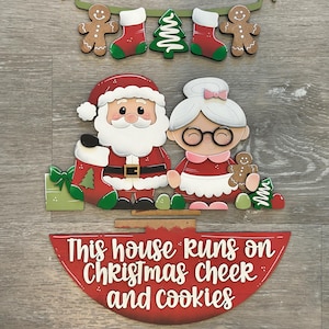 Puede incluir: Decoraci&oacute;n navide&ntilde;a de madera con Pap&aacute; Noel y Mam&aacute; Noel, con el texto "This house runs on Christmas cheer and cookies". El dise&ntilde;o incluye hombres de jengibre, medias y un &aacute;rbol de Navidad.