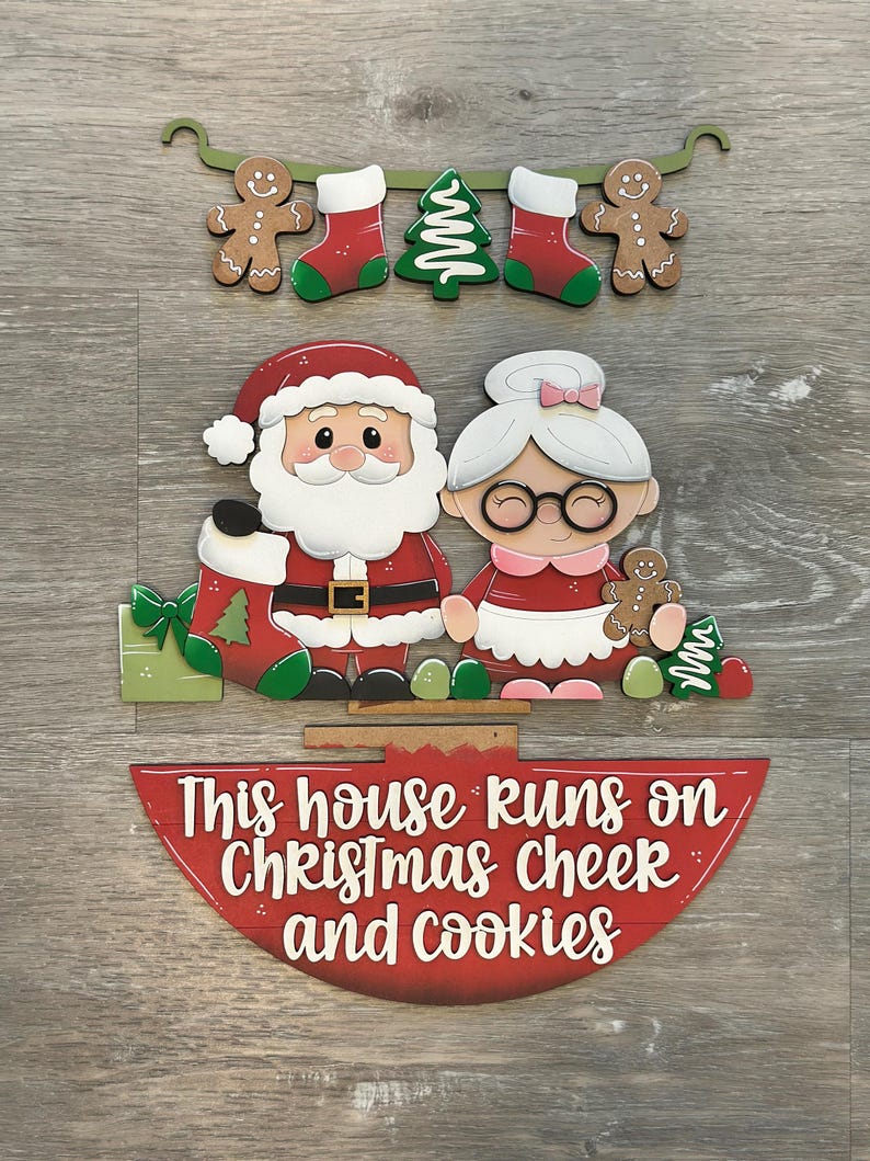 Puede incluir: Decoraci&oacute;n navide&ntilde;a de madera con Pap&aacute; Noel y Mam&aacute; Noel, con el texto "This house runs on Christmas cheer and cookies". El dise&ntilde;o incluye hombres de jengibre, medias y un &aacute;rbol de Navidad.