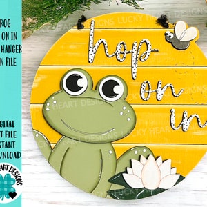 Puede incluir: Un letrero redondo de madera con fondo amarillo y una rana verde con ojos grandes. El letrero dice "hop on in" en letras negras con puntos blancos. Hay un lirio blanco y una pequeña abeja negra y gris en el letrero.
