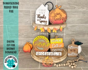 May include: A tiered tray decorated for Thanksgiving with a variety of wooden signs and decorations. The top tier features a white house sign with the text "Let our lives be full of Thanks + Giving", a pumpkin sign with the text "Give Thanks", and a garland of fall leaves. The middle tier features a round wooden sign with the text "Thankful Grateful Blessed", a stack of three signs with the text "Everyone Gather Together", and a small pumpkin. The bottom tier features a wooden sign with the text "Leftovers are for quitters", a wooden sign with the text "Gobble", and a pumpkin.
