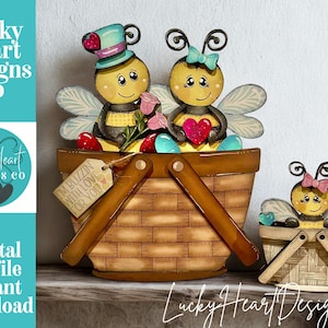 Puede incluir: Un conjunto decorativo de madera con personajes de abejas en cestas de picnic. La cesta principal contiene dos abejas, una con sombrero de copa y otra con lazo, sosteniendo un corazón. El texto "I'm buzzin' with love for you" está en una etiqueta. El conjunto incluye una cesta más pequeña con una sola abeja.