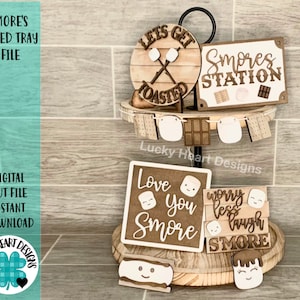 Puede incluir: Una bandeja de tres niveles decorada con un tema de s'mores. El nivel superior tiene un letrero que dice "Let's Get Toasted" con un diseño cruzado de palos para asar malvaviscos. El nivel intermedio tiene un letrero que dice "S'mores Station" y una pancarta con recortes de temática s'mores. El nivel inferior tiene tres letreros que dicen "Love You S'more", "Worry Less Laugh More", y "S'more".