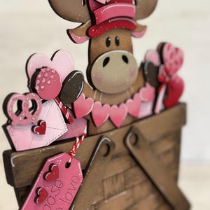 May include: A wooden moose figurine wearing a pink hat and holding a basket filled with pink hearts and other Valentine's Day themed decorations. The basket has a tag that says "I moose be love".