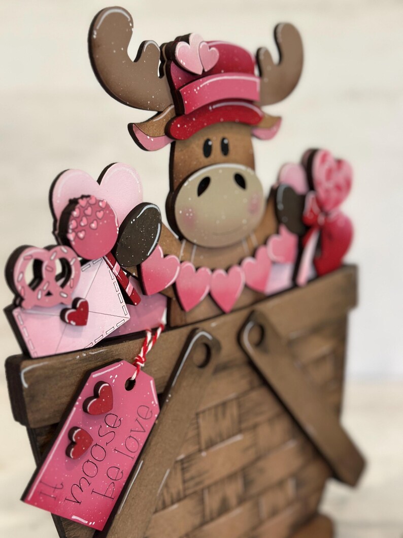 May include: A wooden moose figurine wearing a pink hat and holding a basket filled with pink hearts and other Valentine's Day themed decorations. The basket has a tag that says "I moose be love".