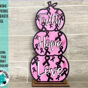 May include: Three pink pumpkins stacked on top of each other. Each pumpkin has a black ribbon design and the words "Faith", "Hope", and "Love" written in white. The pumpkins are on a brown wooden base.