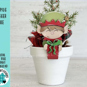 May include: A white ceramic mug hugger with a festive elf design. The elf is wearing a green hat, red and white clothing, and holding a gift. The mug hugger is sitting in a white ceramic pot with a green plant.