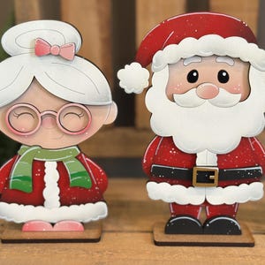 May include: Wooden Christmas decorations featuring Santa Claus and Mrs. Claus. Santa wears a red suit with a white beard and hat. Mrs. Claus has a white hairstyle with a pink bow and glasses. Both figures are on wooden bases.