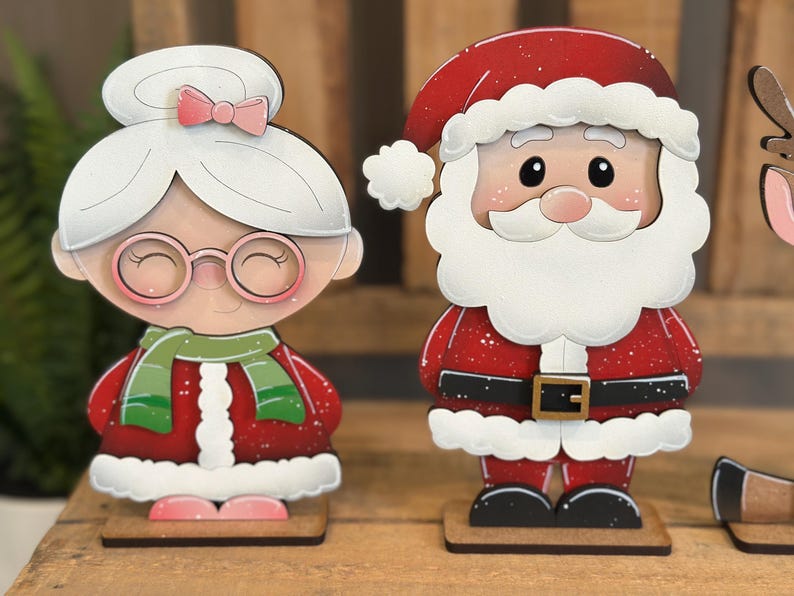 May include: Wooden Christmas decorations featuring Santa Claus and Mrs. Claus. Santa wears a red suit with a white beard and hat. Mrs. Claus has a white hairstyle with a pink bow and glasses. Both figures are on wooden bases.