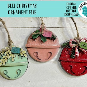 May include: Three wooden Christmas bell ornaments in pastel colors: mint green, coral, and red. Each bell has a jute string for hanging and is decorated with greenery and a tag that says "Merry", "Joy", or "2025".