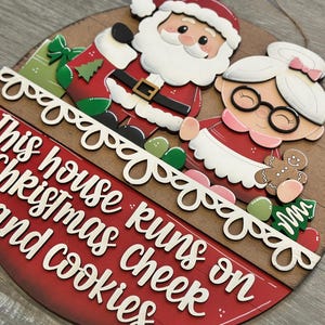 Puede incluir: Un letrero navide&ntilde;o redondo de madera con Santa Claus y la Sra. Claus. El letrero tiene el texto "This house runs on Christmas cheer and cookies" en letras blancas sobre un fondo rojo. Incluye un hombre de jengibre y un &aacute;rbol de Navidad. Aproximadamente 25 cm de di&aacute;metro.