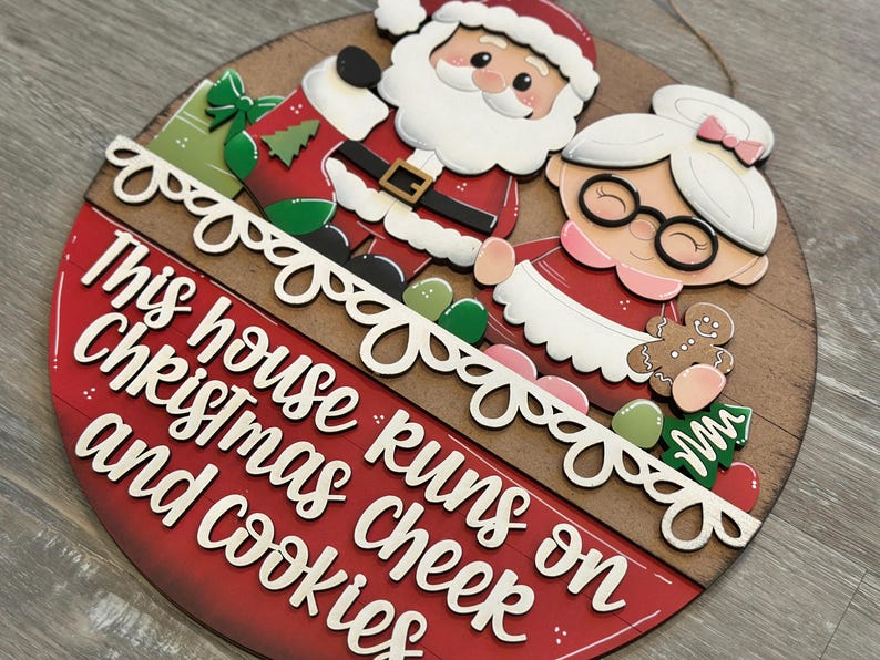 Puede incluir: Un letrero navide&ntilde;o redondo de madera con Santa Claus y la Sra. Claus. El letrero tiene el texto "This house runs on Christmas cheer and cookies" en letras blancas sobre un fondo rojo. Incluye un hombre de jengibre y un &aacute;rbol de Navidad. Aproximadamente 25 cm de di&aacute;metro.